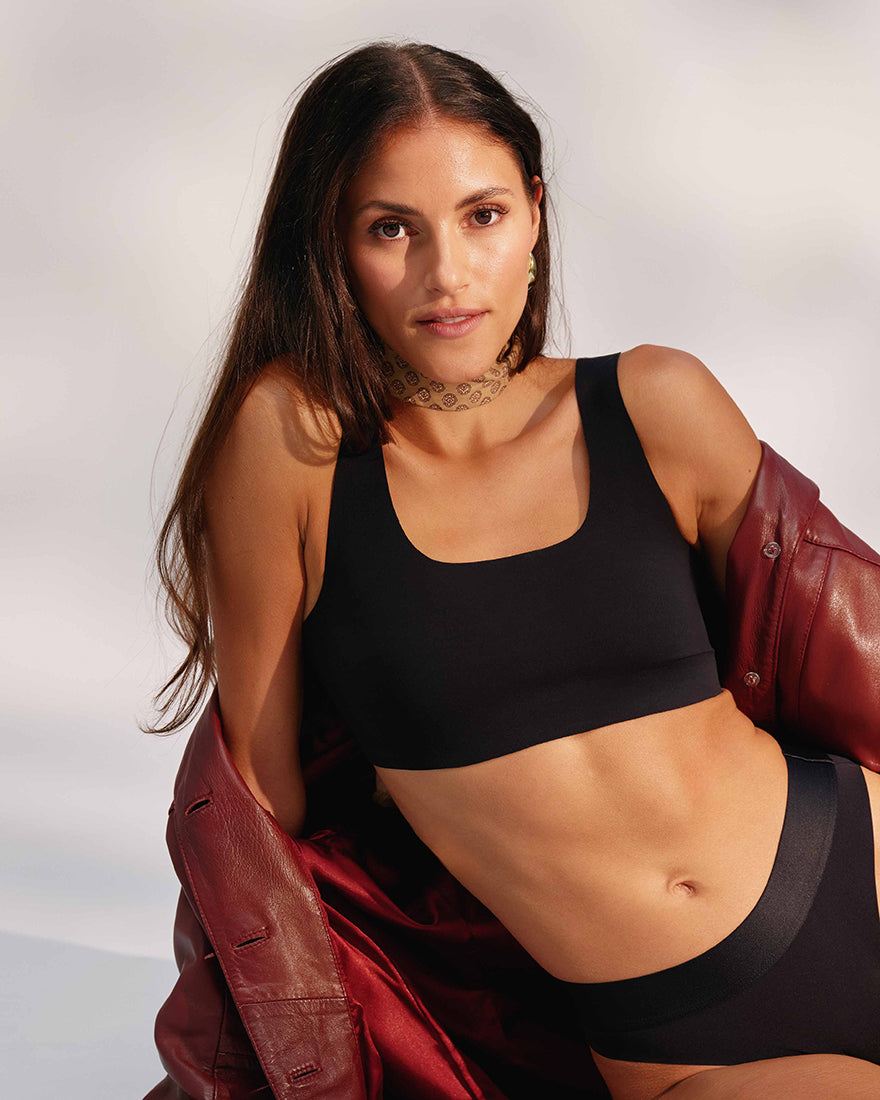 Modell trägt ooia All-You-Need Bra Top in Schwarz – bügelloses, nahtloses Bustier mit breiten Trägern, mit roter Jacke und ooia Slip Cheeky in Schwarz.