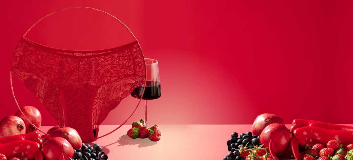 Der Periodenslip Hipster Allover Lace in Rot steht auf einem Tisch mit Rotwein, Erdbeeren, Äpfeln und weiterem roten Obst und Gemüse.
