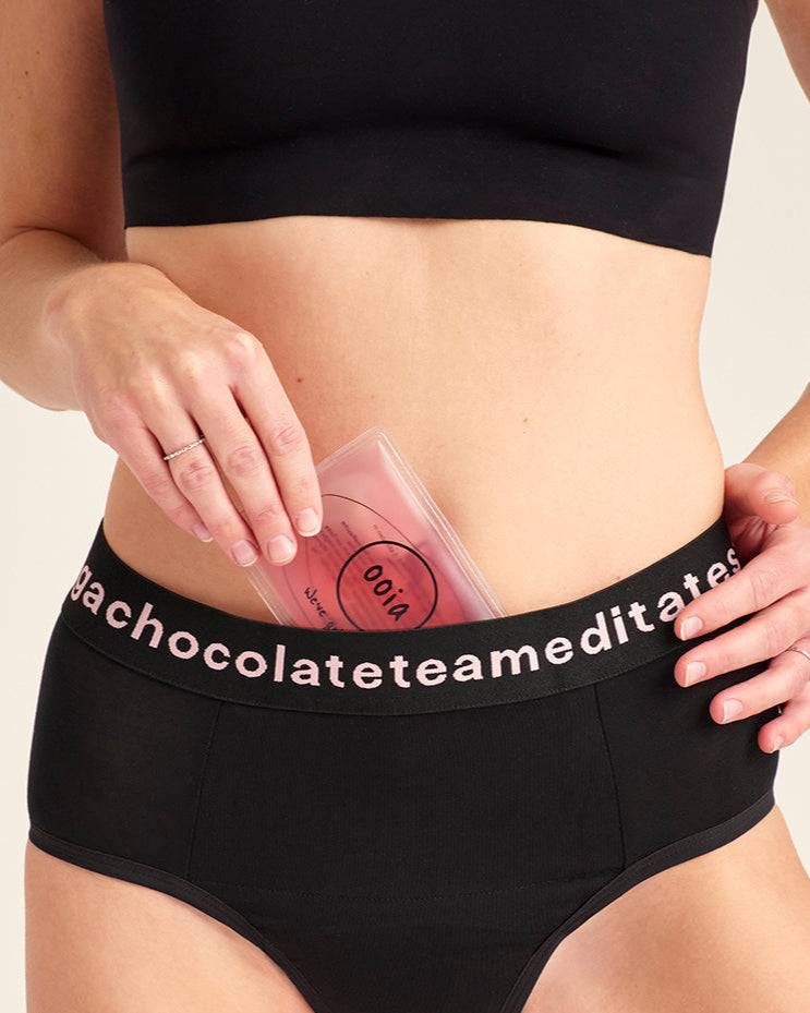 Modell trägt ooia Slip Hugs in Schwarz – bequemer Slip mit extra stützendem Bund mit Aufdruck und integrierten Taschen für das ooia Wärmepad. Model tut das Wärmepad in die vordere Tasche.