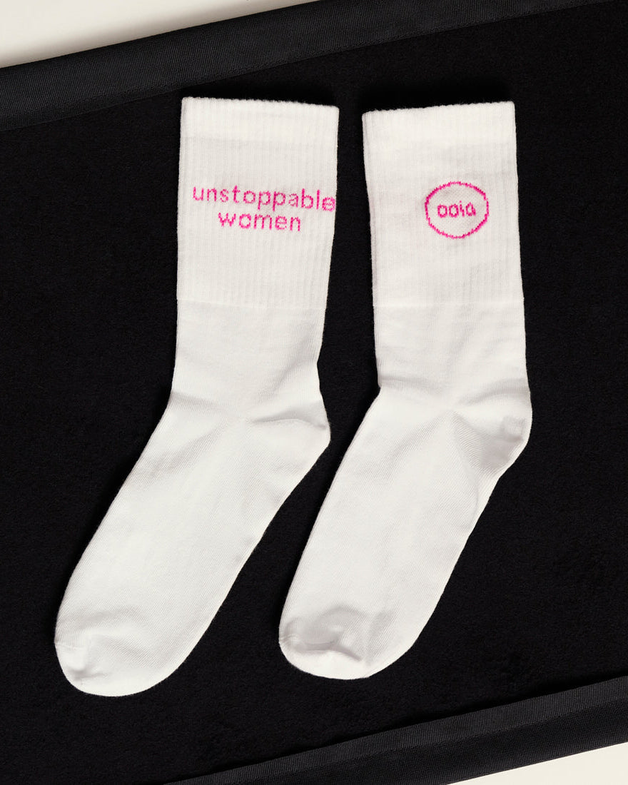 weiße ooia Socken mit dem pinken Aufdruck: Unstoppable Woman, liegen auf einer dunklen Fläche 