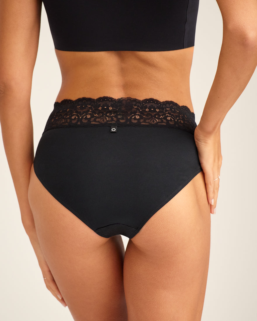 Modell trägt ooia everyday Slip in Schwarz – Unterwäsche mit breitem Spitzenbund, die Slipeinlagen ersetzt. Rückansicht.